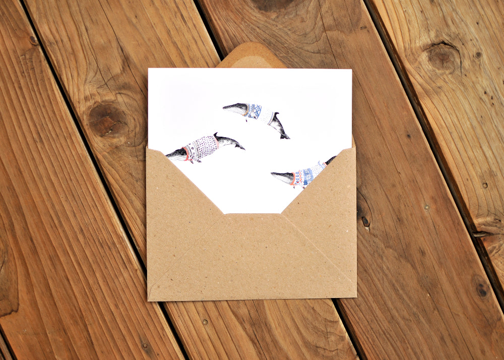 Greeting card with three minke whales wearing Norwegian ski sweaters, called lusekoftes. Card is partway in an envelope.