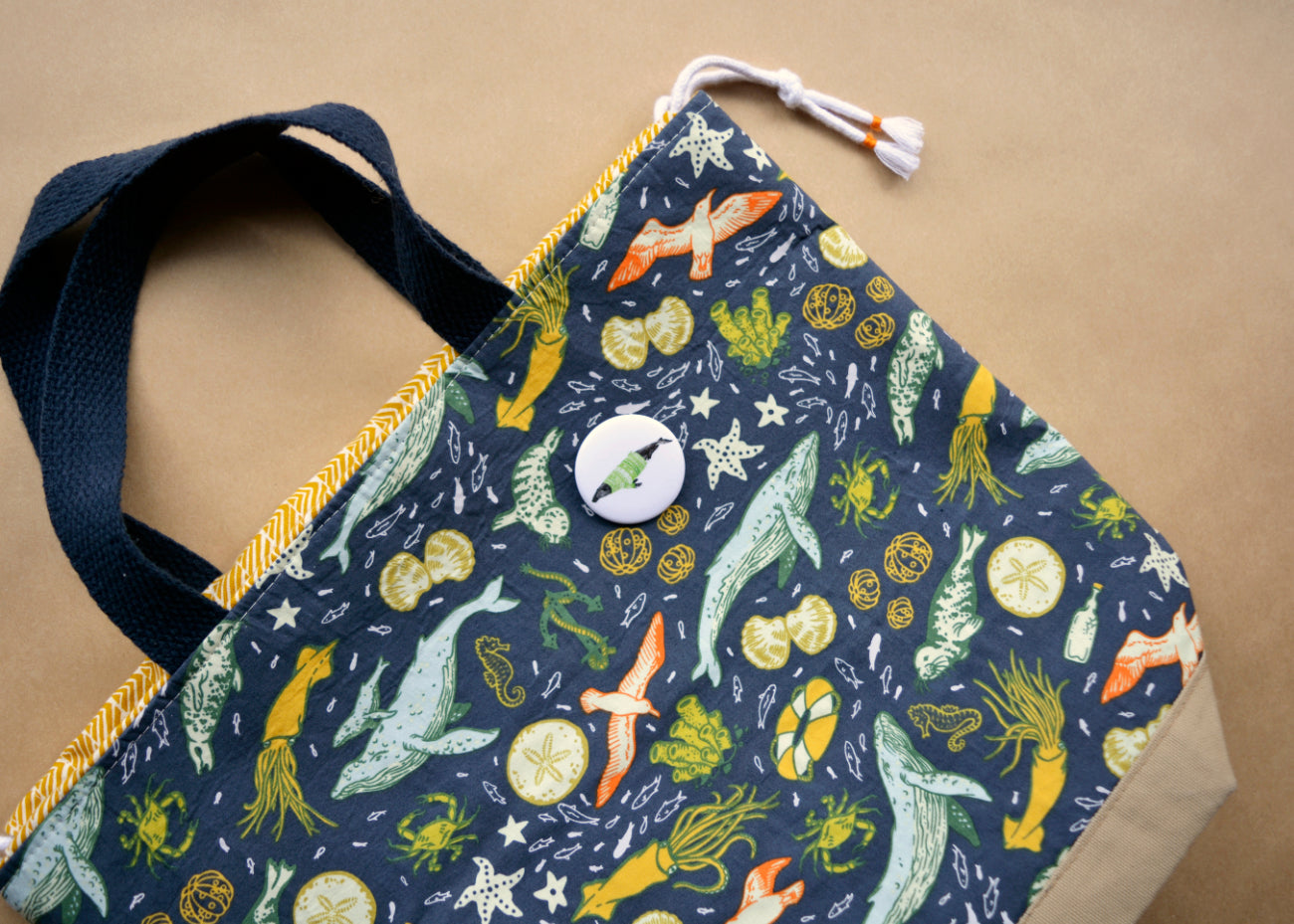 Ink drawing of a humpback whale wearing a green sweater on a pin-backed button on a cloth bag.