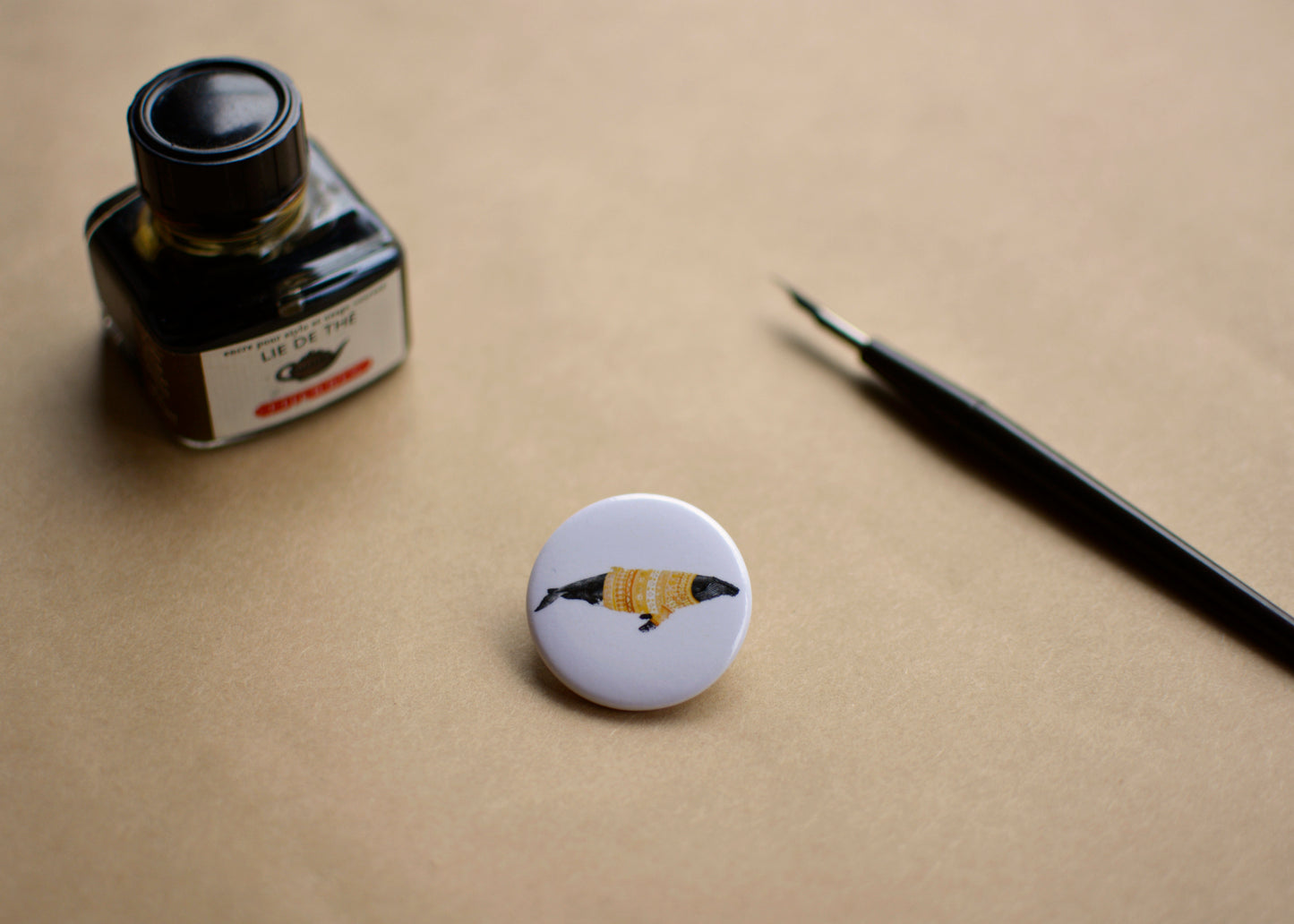 Ink drawing of a humpback whale wearing a green sweater on a pin-backed button. Image features a ink bottle and a dip pen.