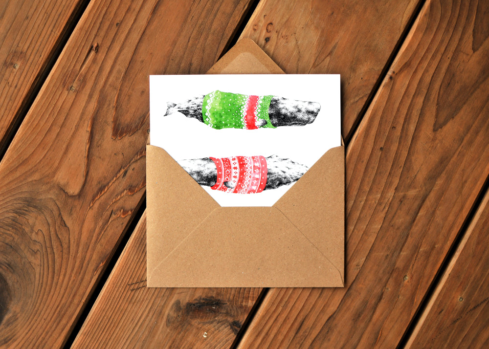 A card with a ink dawing of a humpback whale wearing a colorful sweater. Card is partway out of an envelope.