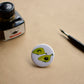 Pin back button with two imaginative "Avocado whales", next to ink bottle and dip pen.