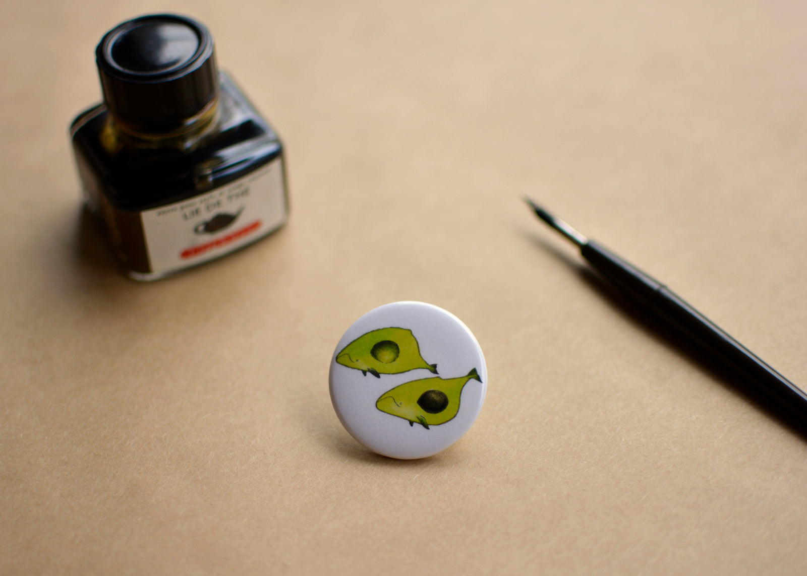 Pin back button with two imaginative "Avocado whales", next to ink bottle and dip pen.