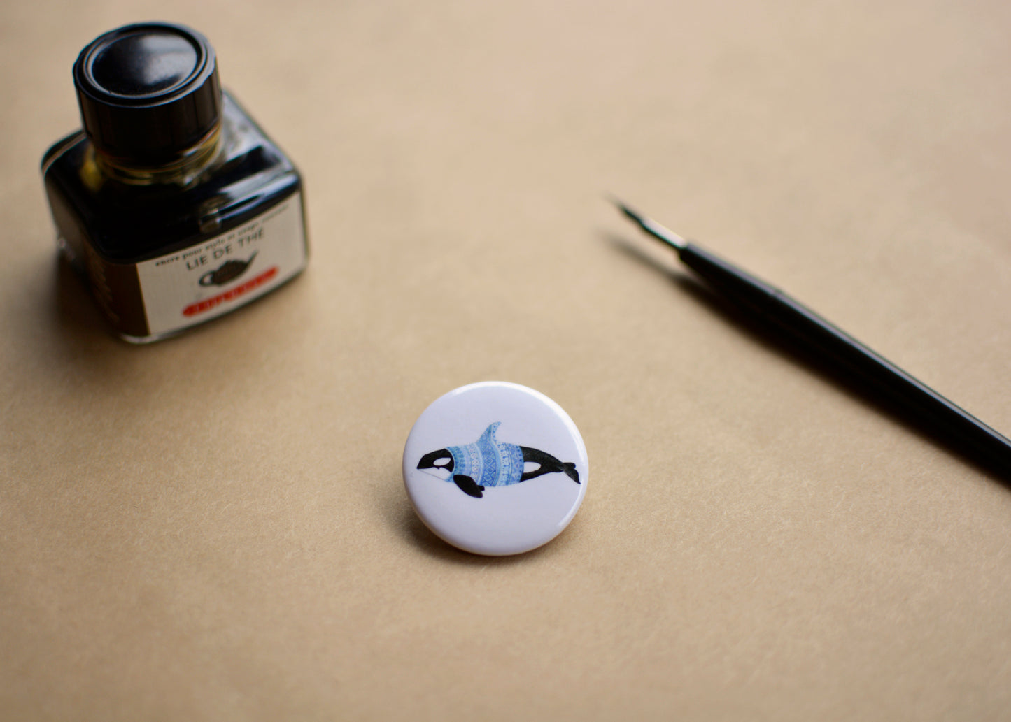 Ink drawing of a humpback whale wearing a blue sweater on a pin-backed button. Ink bottle and dip pen shown with the button.