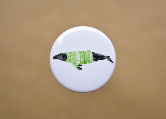 Ink drawing of a humpback whale wearing a green sweater on a pin-backed button.