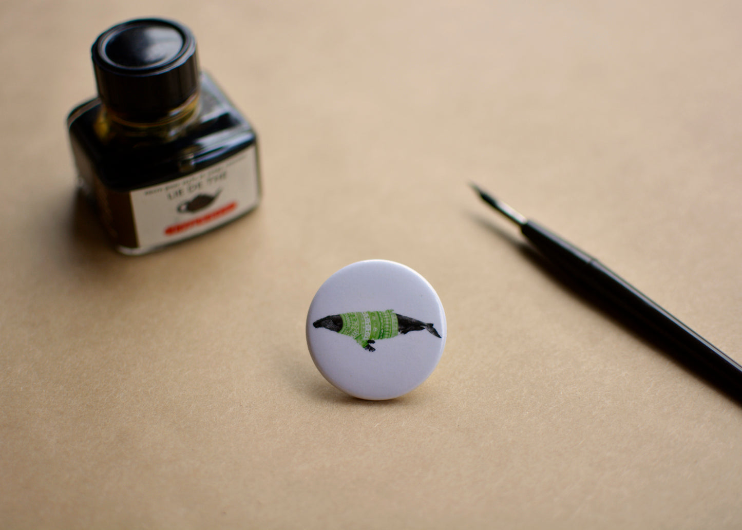 Ink drawing of a humpback whale wearing a green sweater on a pin-backed button. Image features a ink bottle and dip pen.