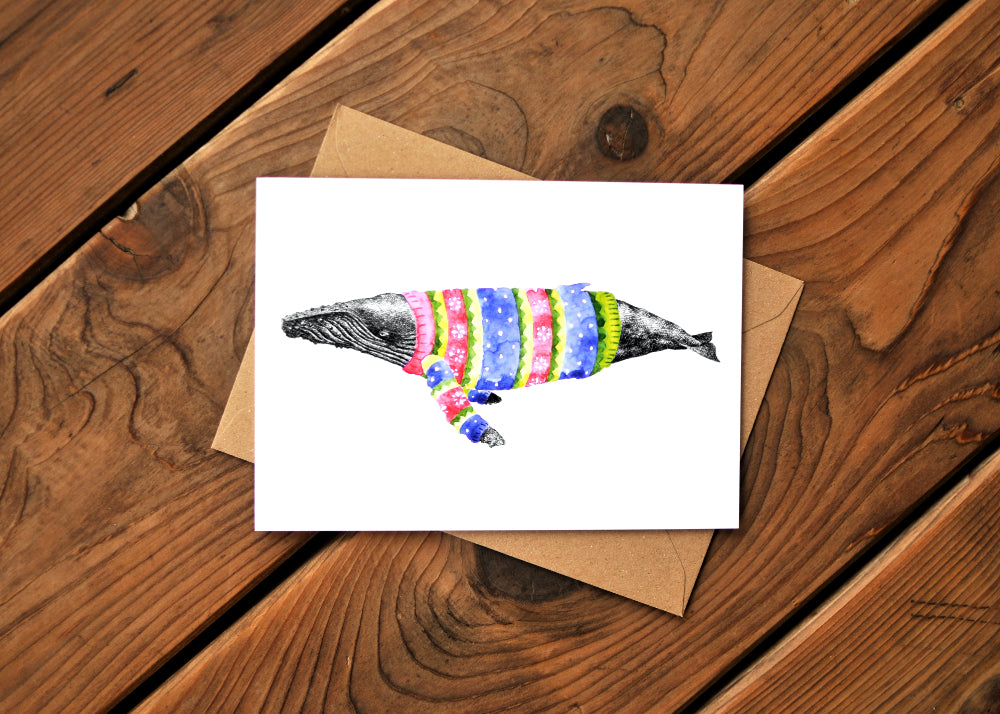A card with a ink dawing of a humpback whale wearing a colorful sweater. Card is on top of an envelope.