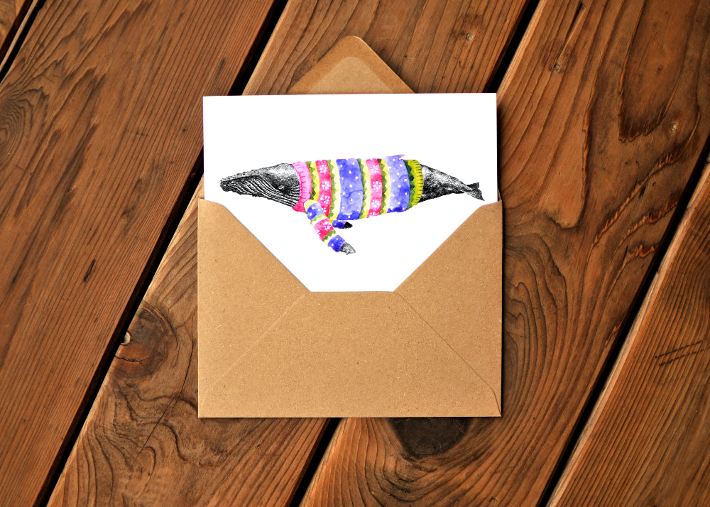 A card with a ink dawing of a humpback whale wearing a colorful sweater. Card in partway in an envelope.