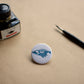 Pin back button with an imaginative skeleton whale. Ink bottle and dip pen featured with the pin.