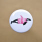 Ink drawing of a humpback whale wearing a pink sweater on a pin-backed button.