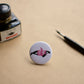 Ink drawing of a humpback whale wearing a pink sweater on a pin-backed button. Featured with a ink bottle and dip pen.