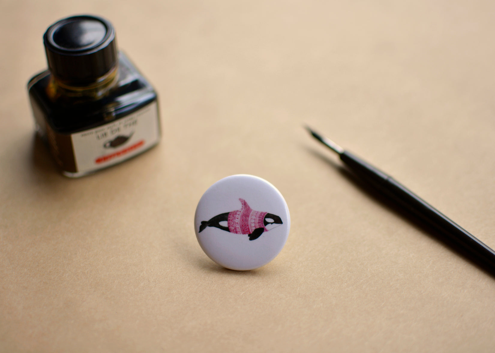 Ink drawing of a humpback whale wearing a pink sweater on a pin-backed button. Featured with a ink bottle and dip pen.
